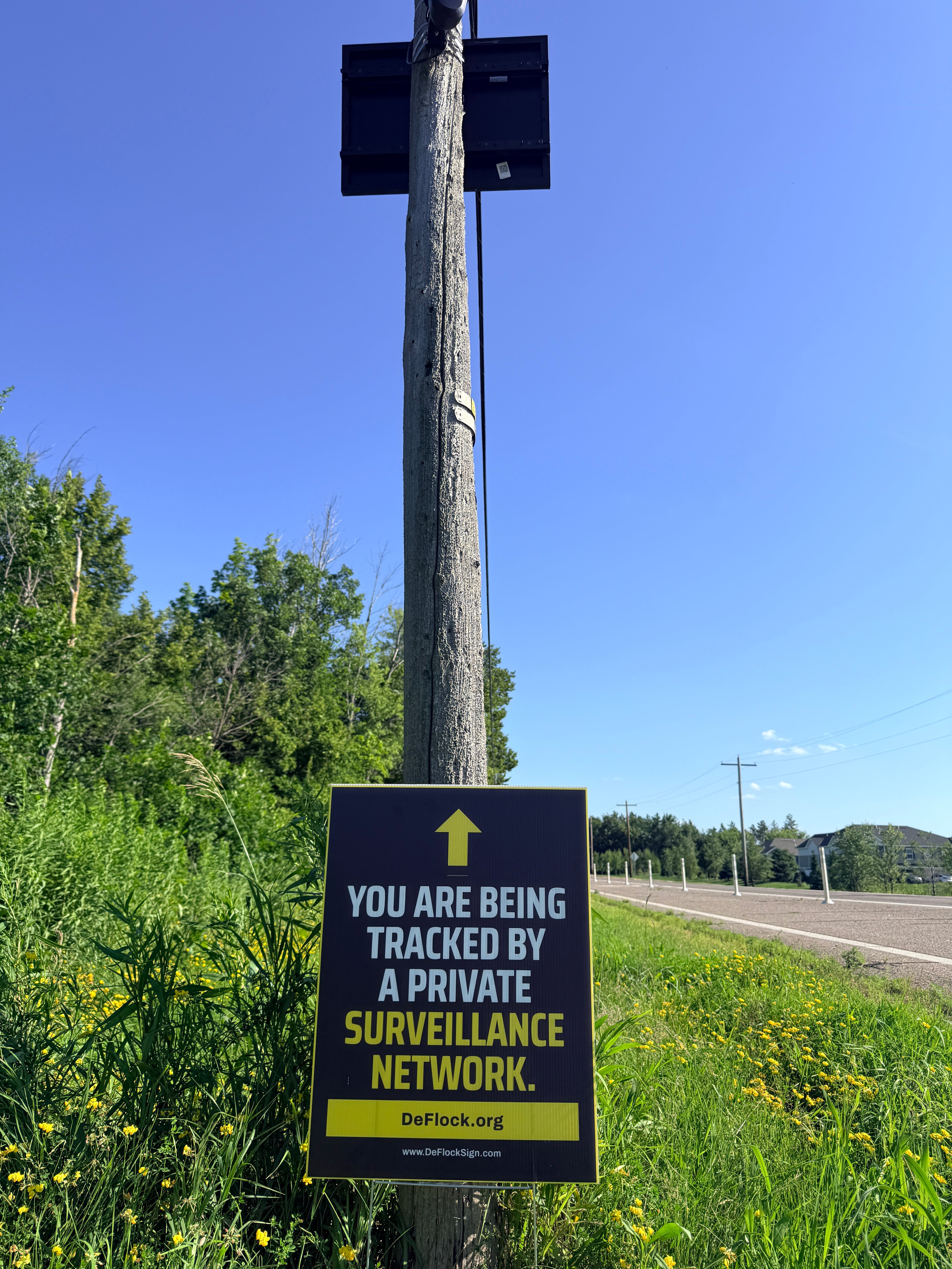 Yard sign below a Flock camera with an arrow pointing upwards, and a message that says "You are being tracked by a private surveillance network" to indicate that above the sign is a surveillance camera.