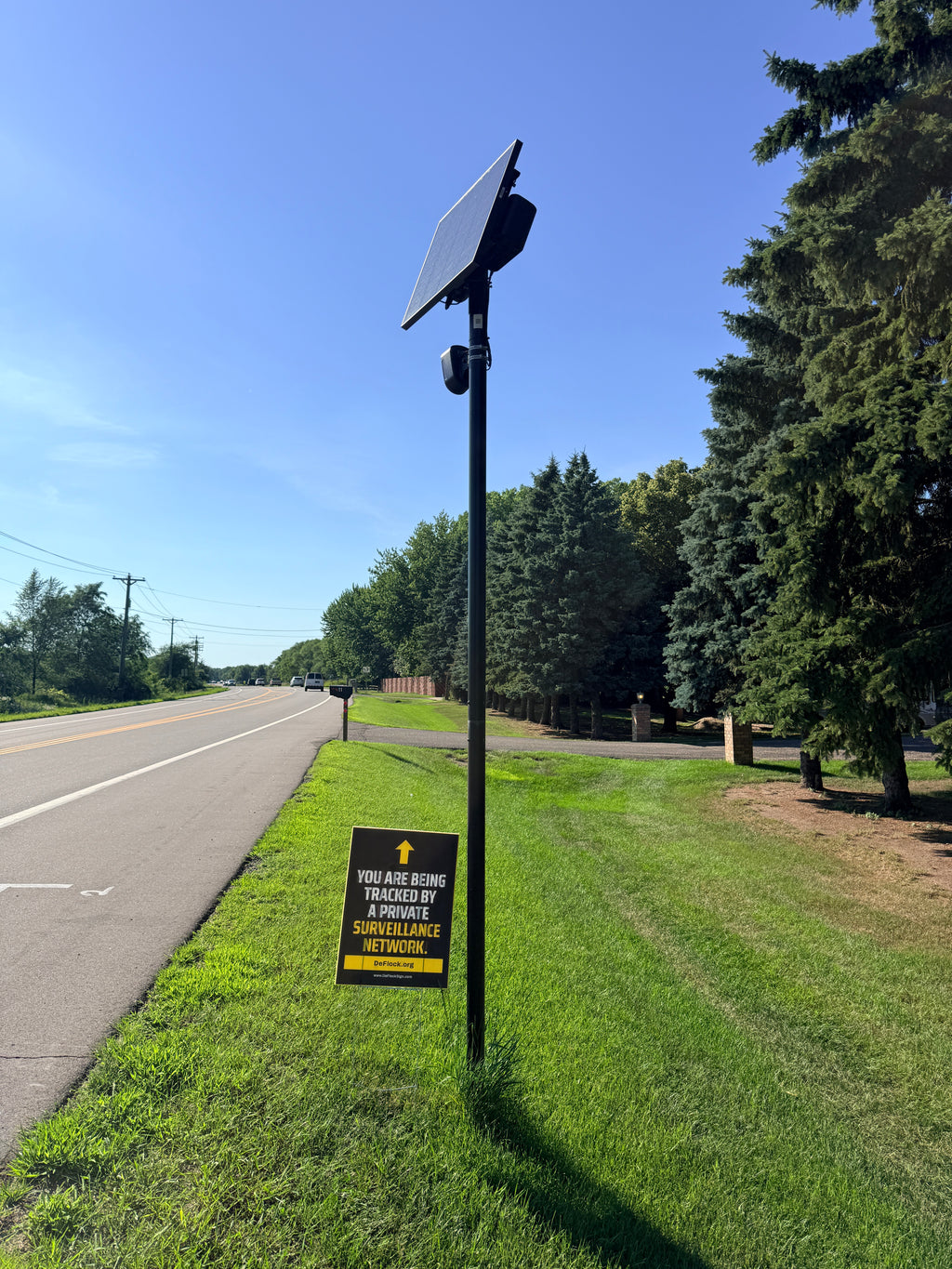 Yard sign below a Flock camera with an arrow pointing upwards, and a message that says "You are being tracked by a private surveillance network" to indicate that above the sign is a surveillance camera.
