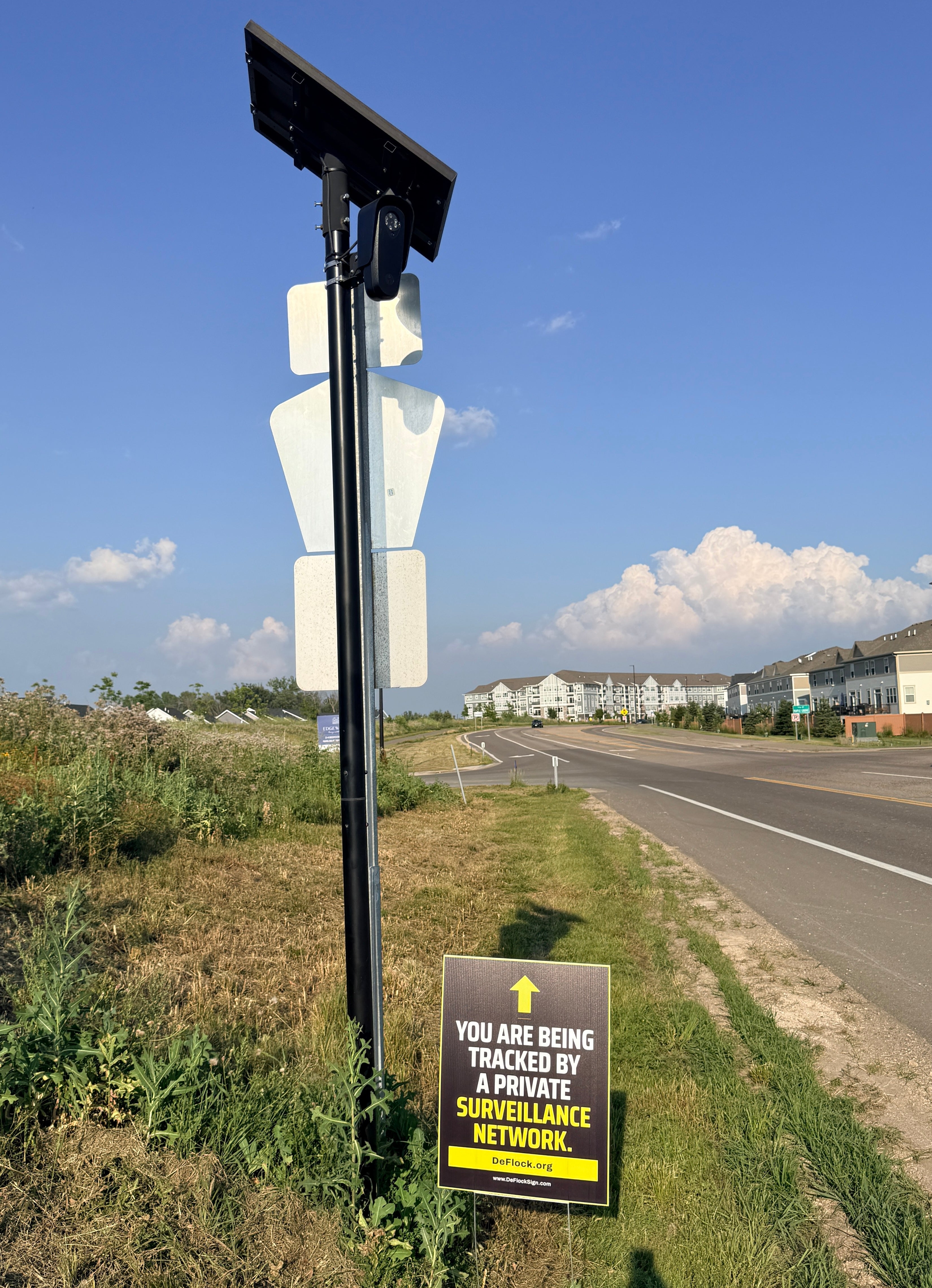 Yard sign below a Flock camera with an arrow pointing upwards, and a message that says "You are being tracked by a private surveillance network" to indicate that above the sign is a surveillance camera.