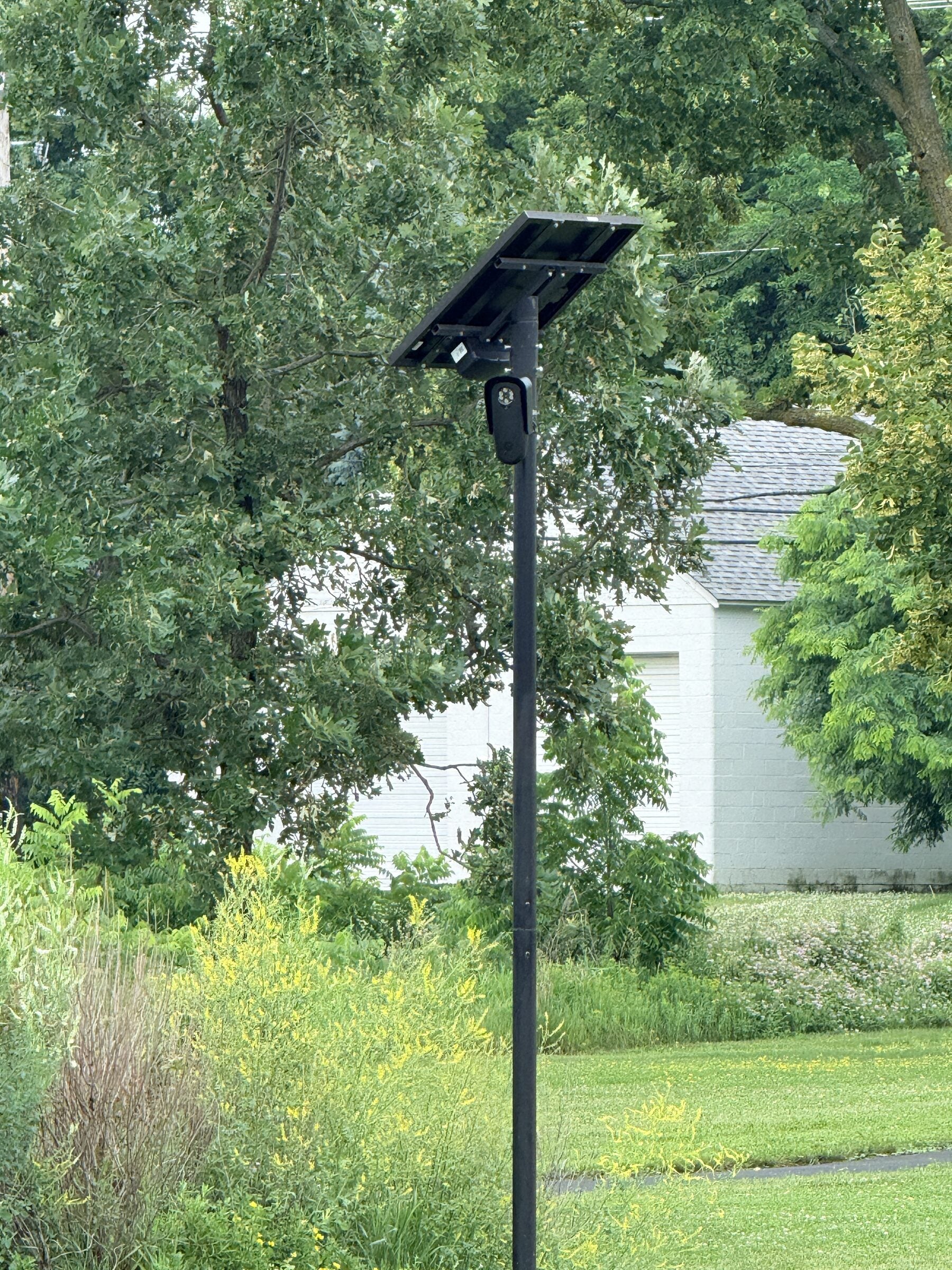 A Flock automated license plate reader camera on a black pole against trees and a farmhouse