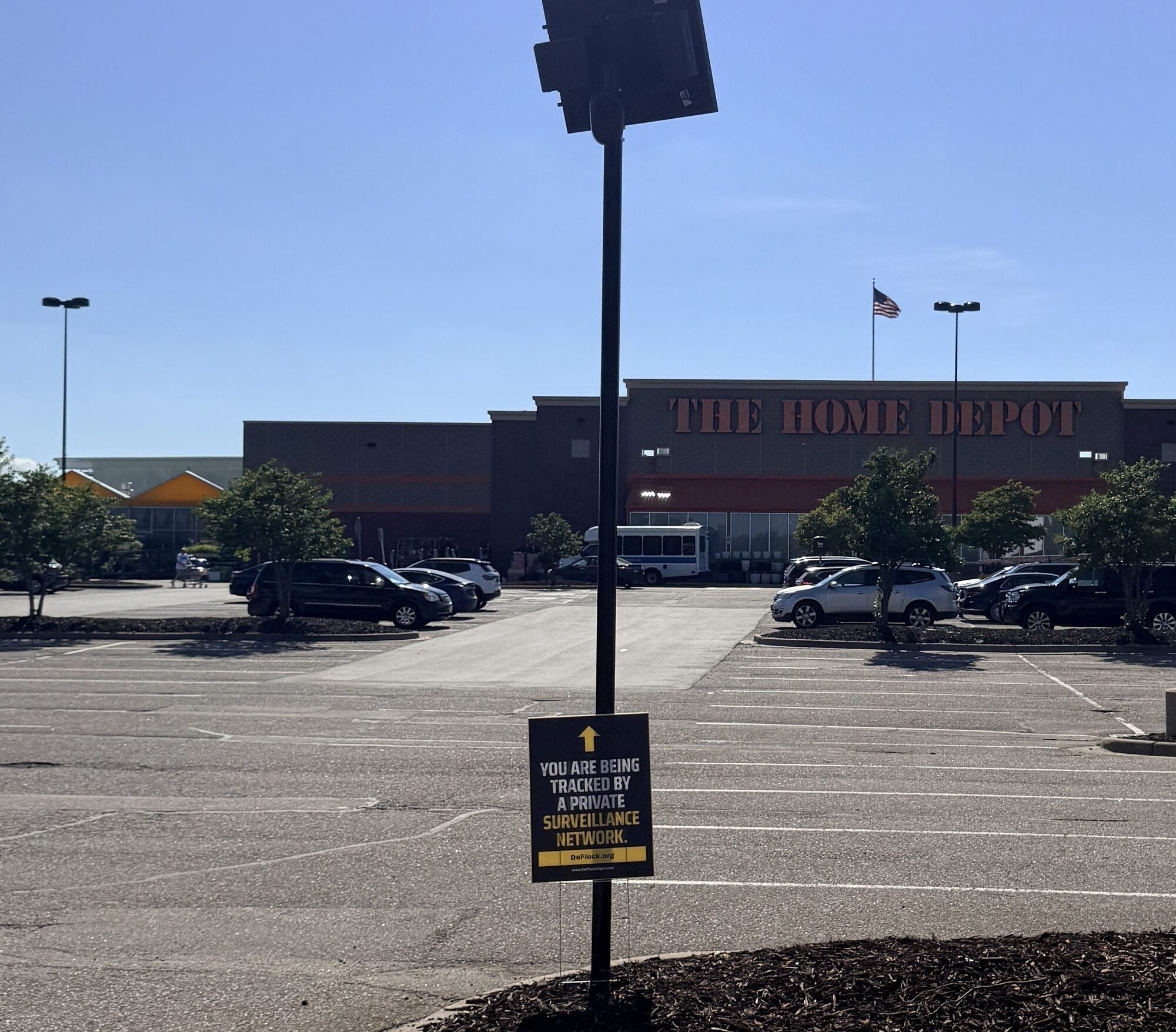 Official DeFlock sign beneath a pole-mounted automated license plate reader in a Home Depot parking lot