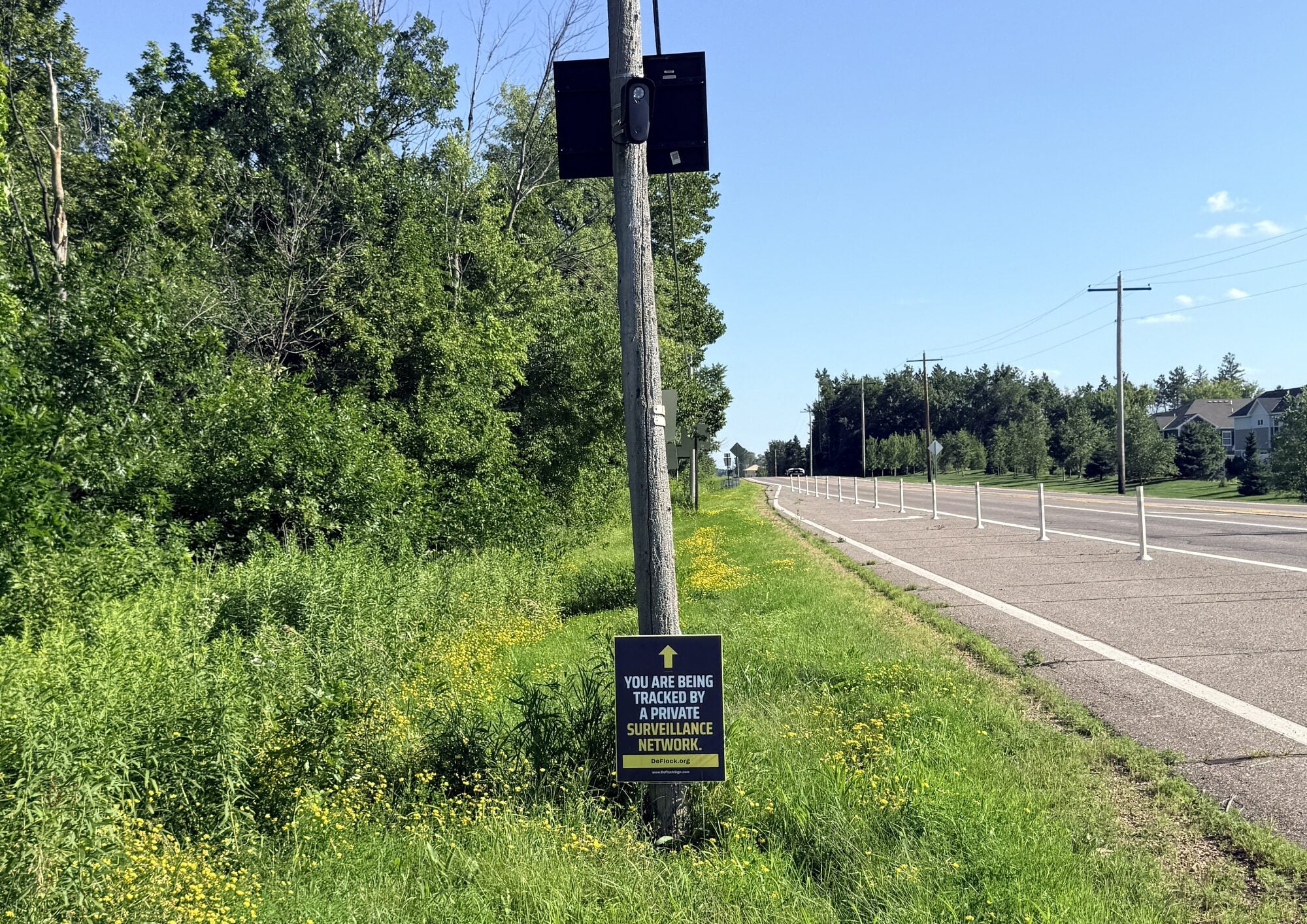 Official DeFlock sign mounted below an automated license plate reader on a wooden utility pole beside a road