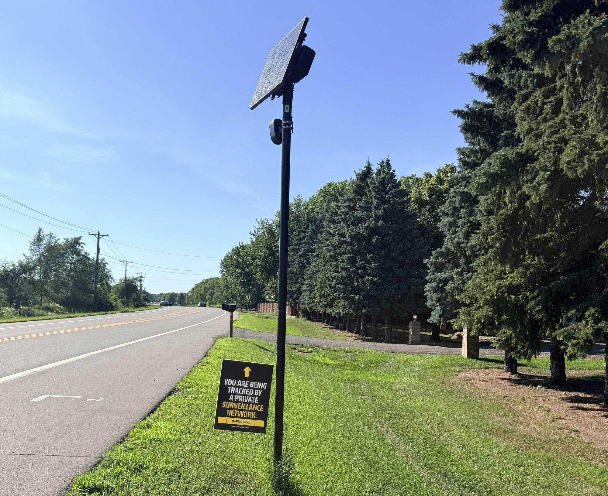 Official DeFlock sign at the base of a solar-powered automated license plate reader beside a neighborhood road